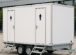 Een witte mobiele toiletwagen voor 8 personen met aparte deuren voor dames en heren, geplaatst op een evenementenlocatie